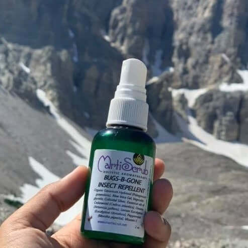 Hand holding Bugs-B-Gone insect repellent spray in a mountain landscape, featuring organic ingredients for outdoor comfort.