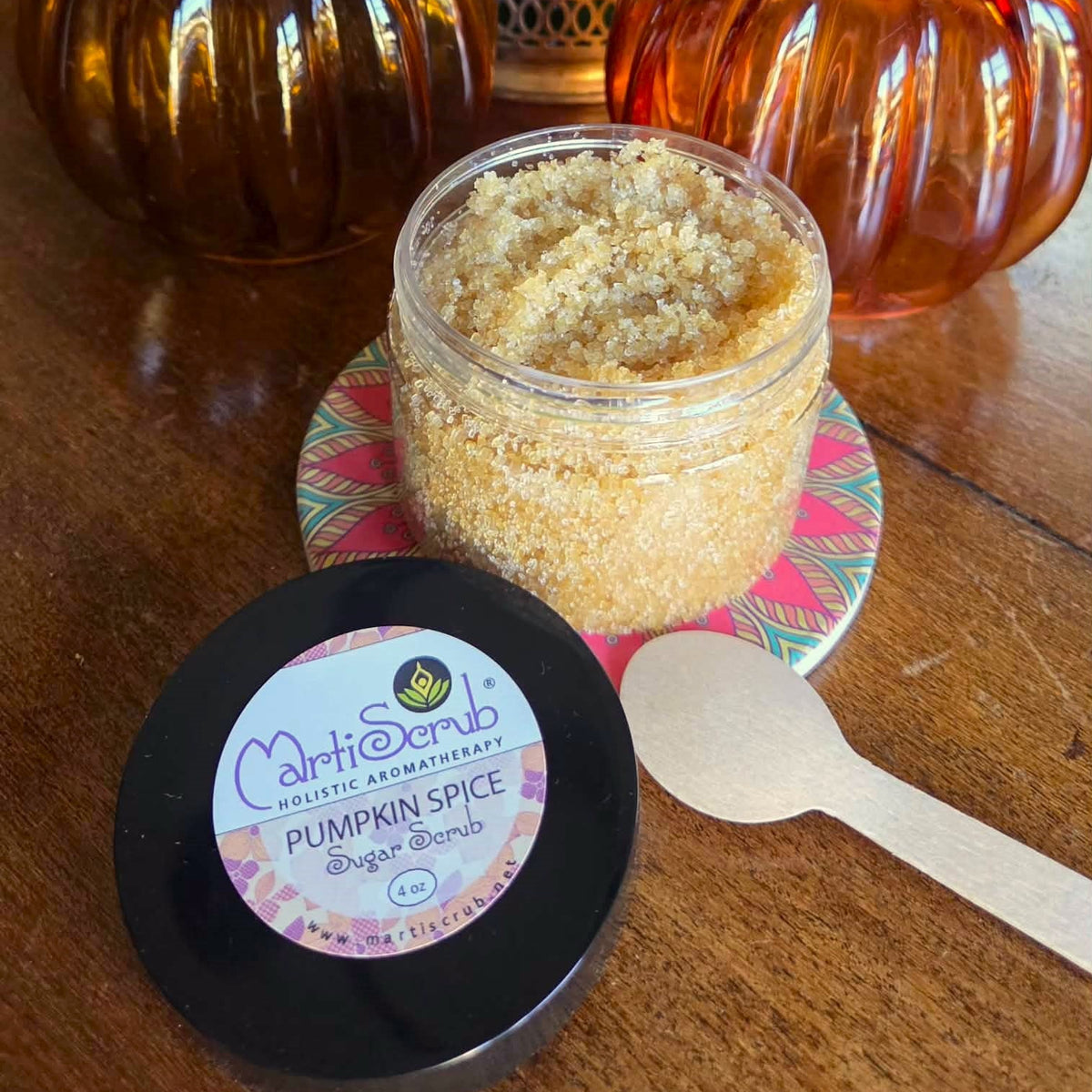 A jar of CarrieSunclo Holistic Remedies Pumpkin Spice Body Scrub sits on a patterned coaster on a wooden table, with its black lid in the foreground and decorative amber glass pumpkins in the background.